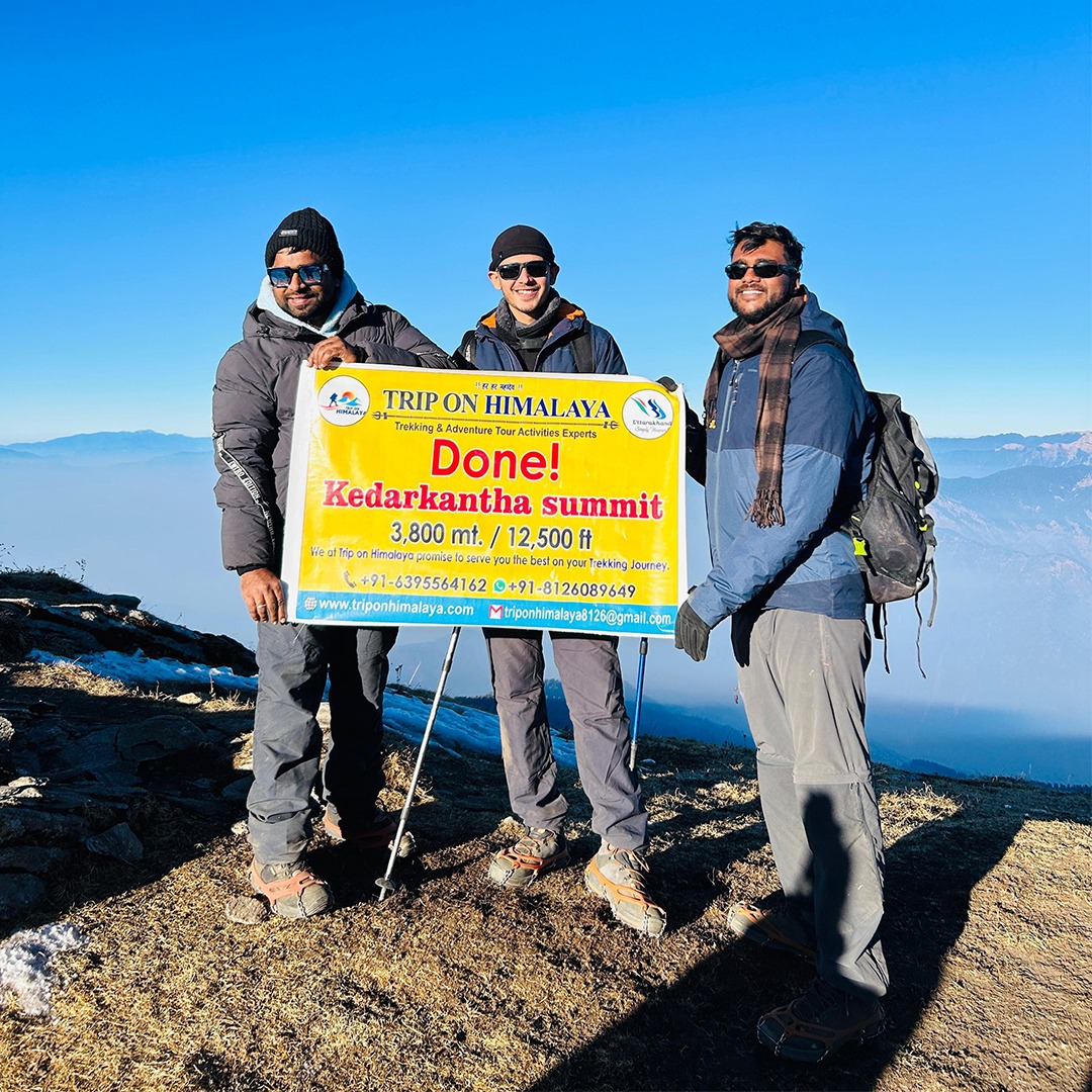 TripOnHimalaya banner held at Kedarkantha Summit with snow-covered Himalayan peaks in the background
