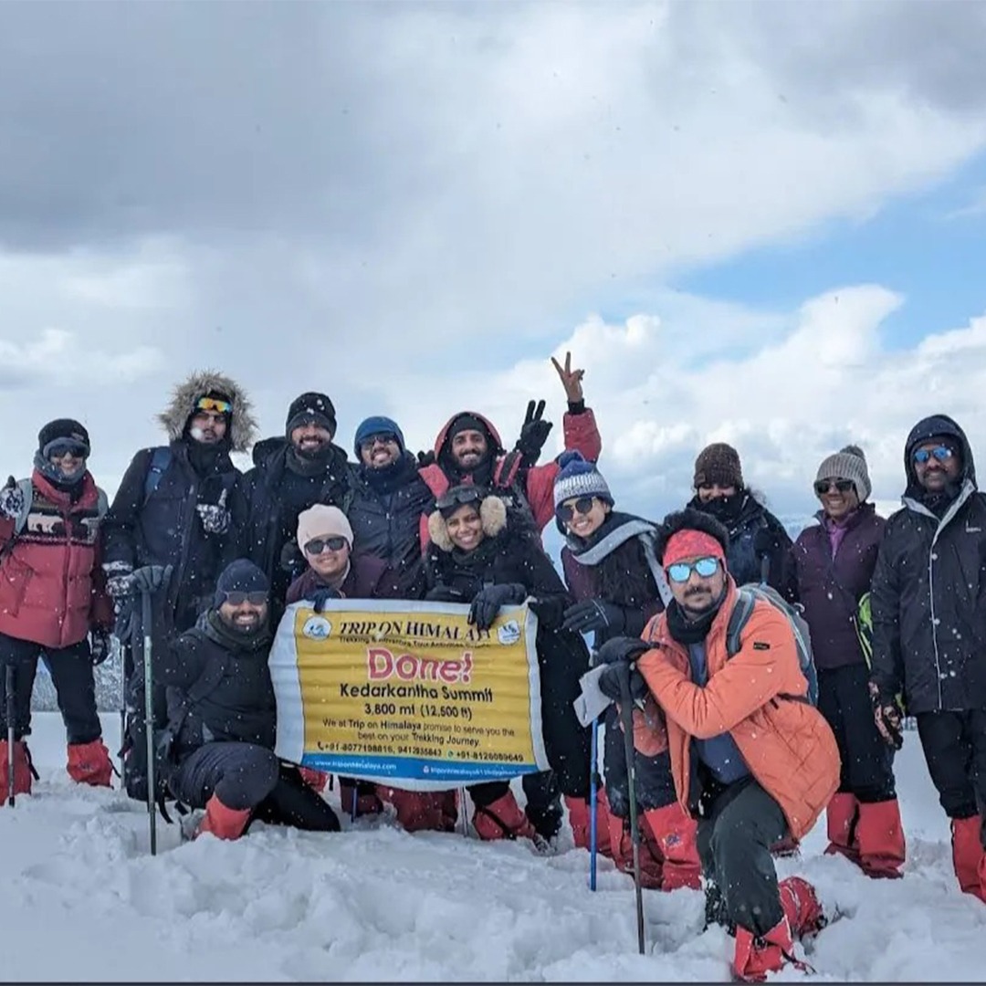 Trekkers holding a celebratory banner at Kedarkantha Summit against a snowy backdrop